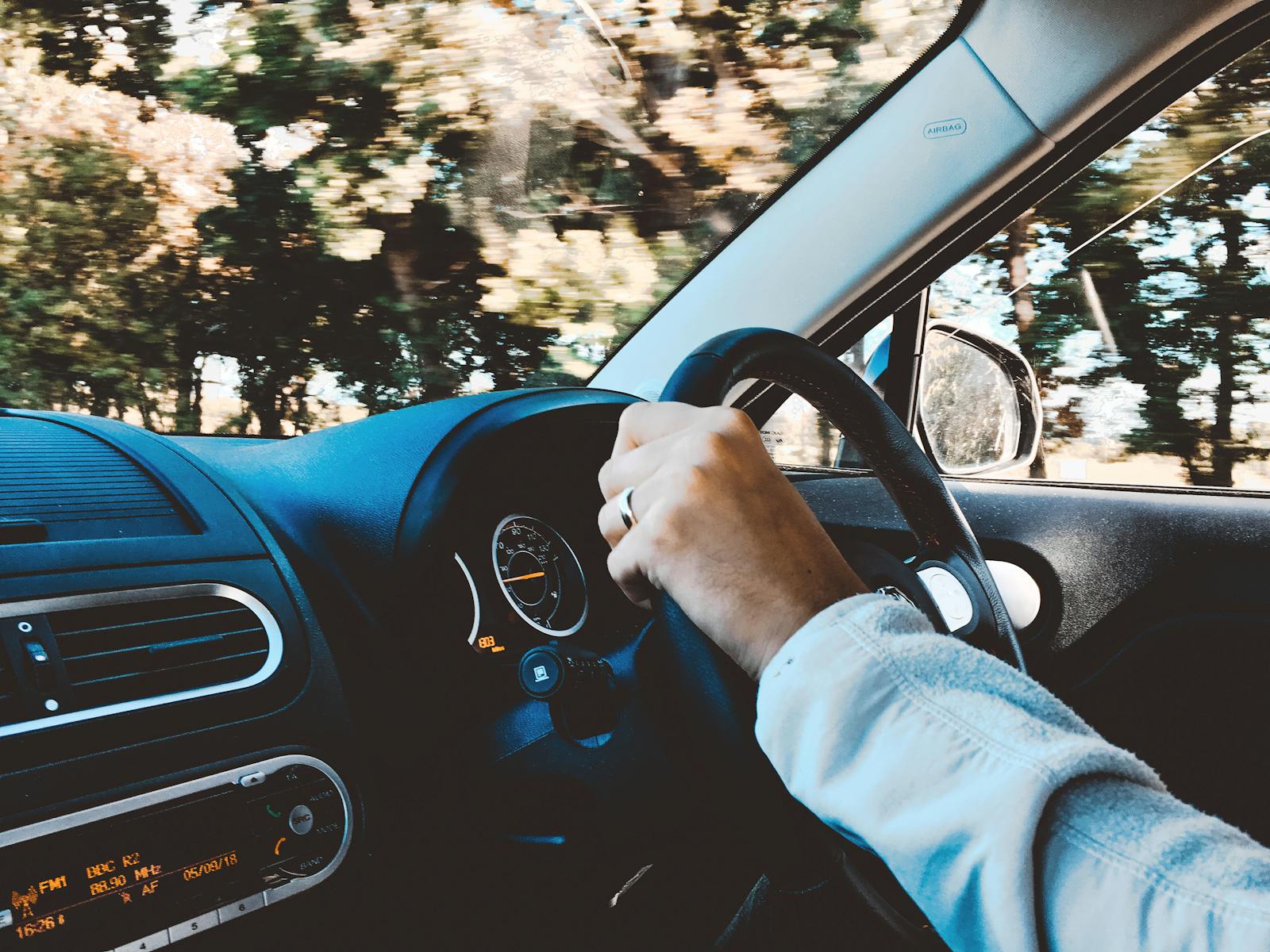 Driver's hand on steering wheel of a modern car during a daytime drive through scenic outdoors.